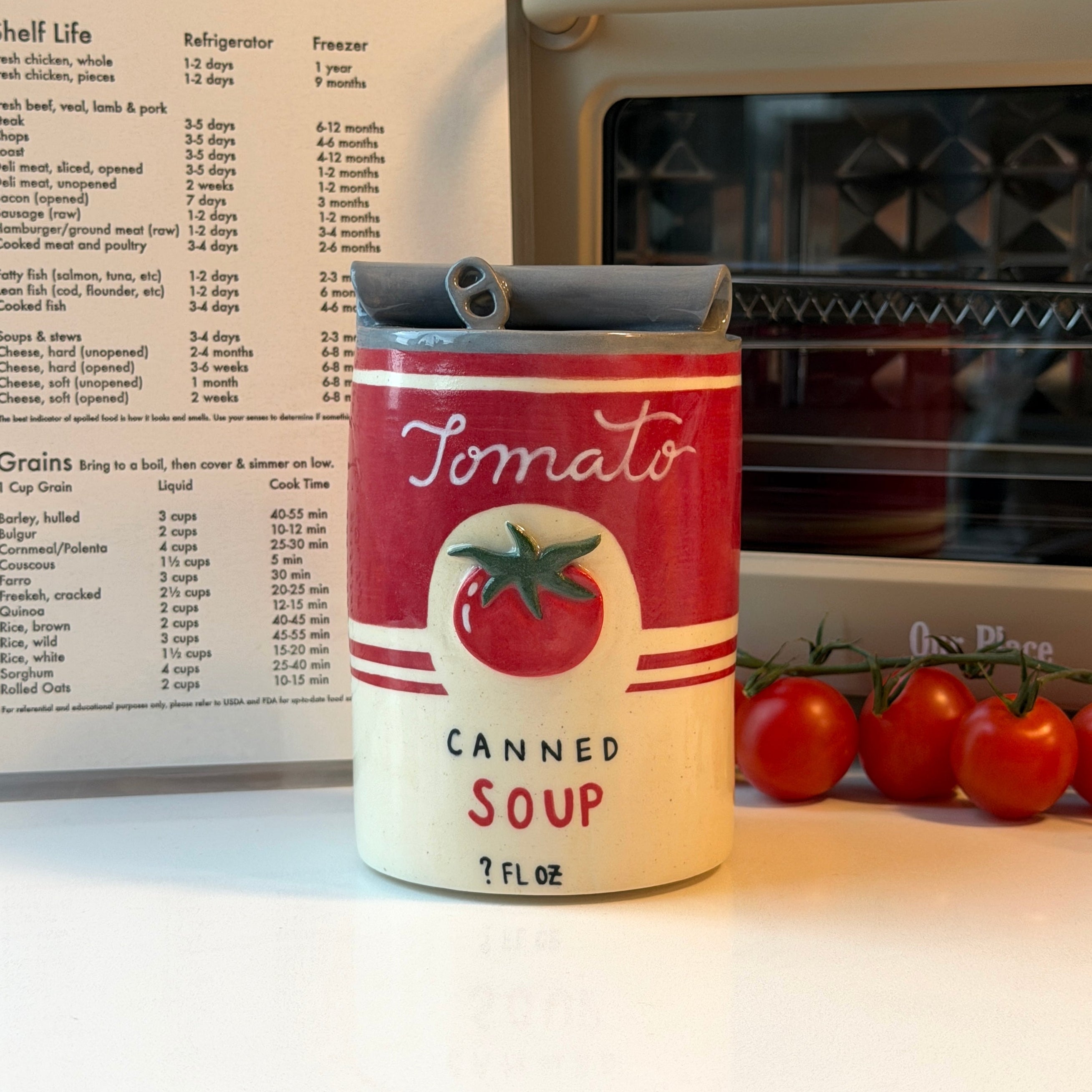 A novelty can-shaped ceramic mug that looks like tomato soup sits on a kitchen counter in front of an oven and recipe card, with fresh tomatoes beside it.