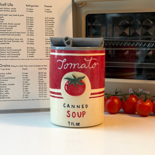 A novelty can-shaped ceramic mug that looks like tomato soup sits on a kitchen counter in front of an oven and recipe card, with fresh tomatoes beside it.