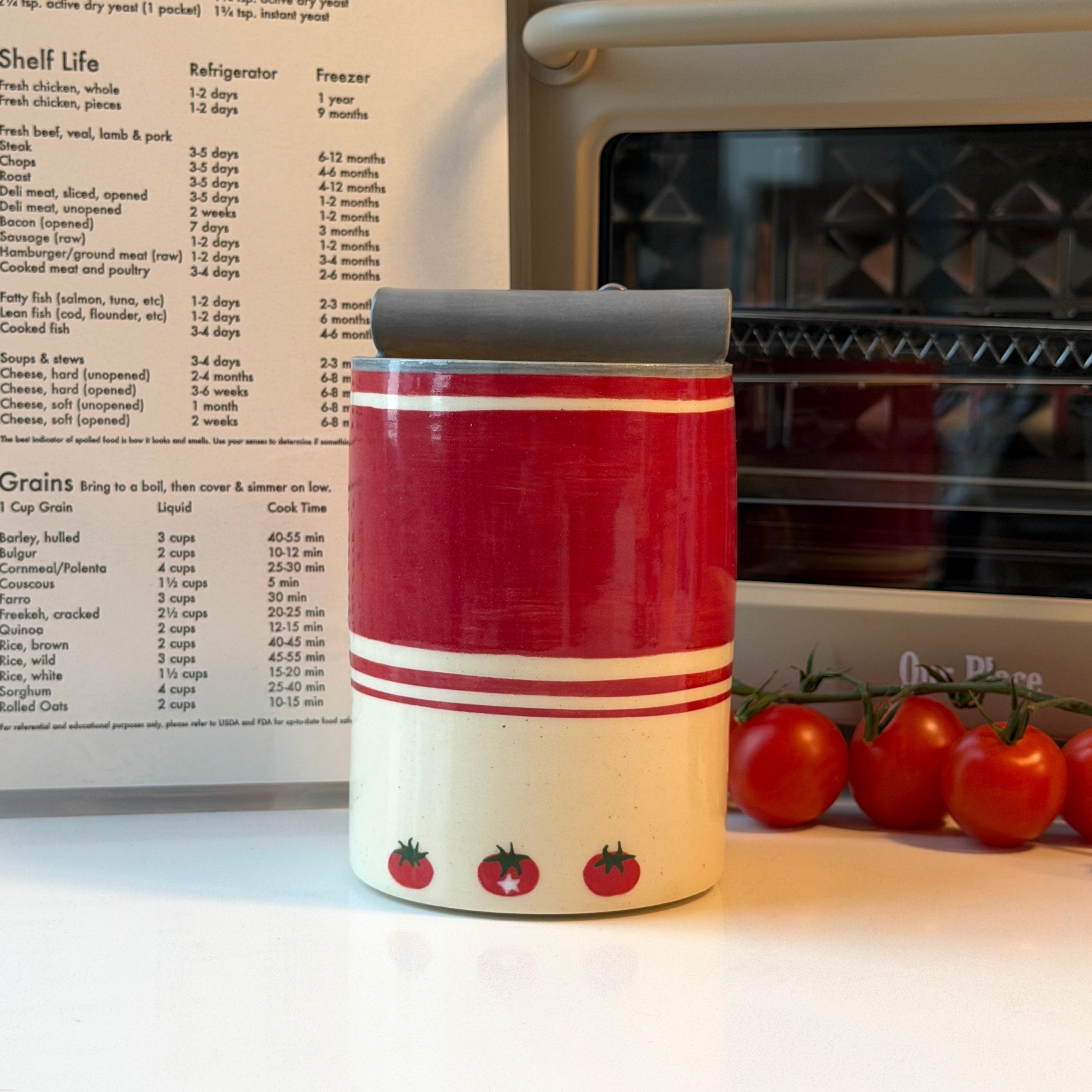 A red and cream ceramic mug with a dark gray lid sits on a white countertop in a kitchen setting, with a recipe book, oven, and tomatoes in the background.