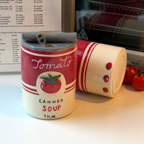 Two ceramic can-shaped mugs styled like retro tomato soup cans, one standing upright and the other lying on its side, placed on a white countertop with tomatoes and a stove in the background.