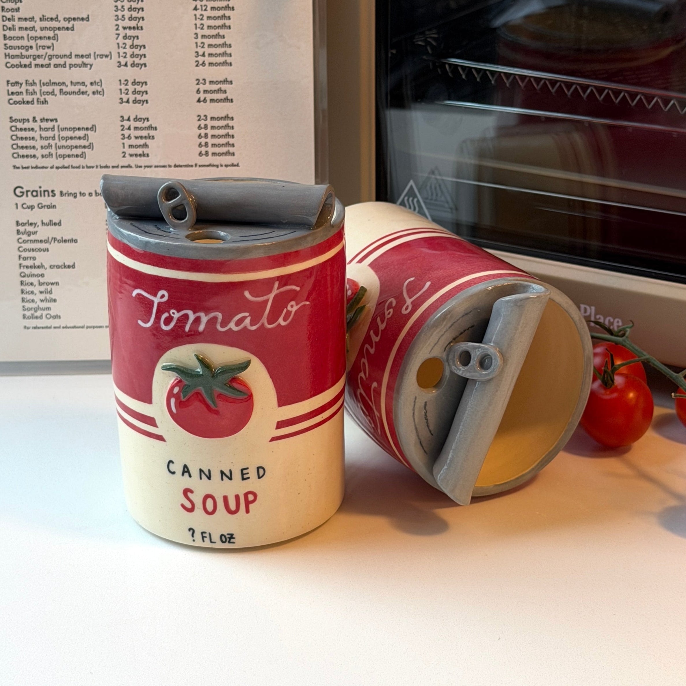 Two novelty ceramic mugs styled like vintage tomato soup cans, one standing upright and the other lying on its side, on a white countertop with a recipe sheet and an oven in the background.