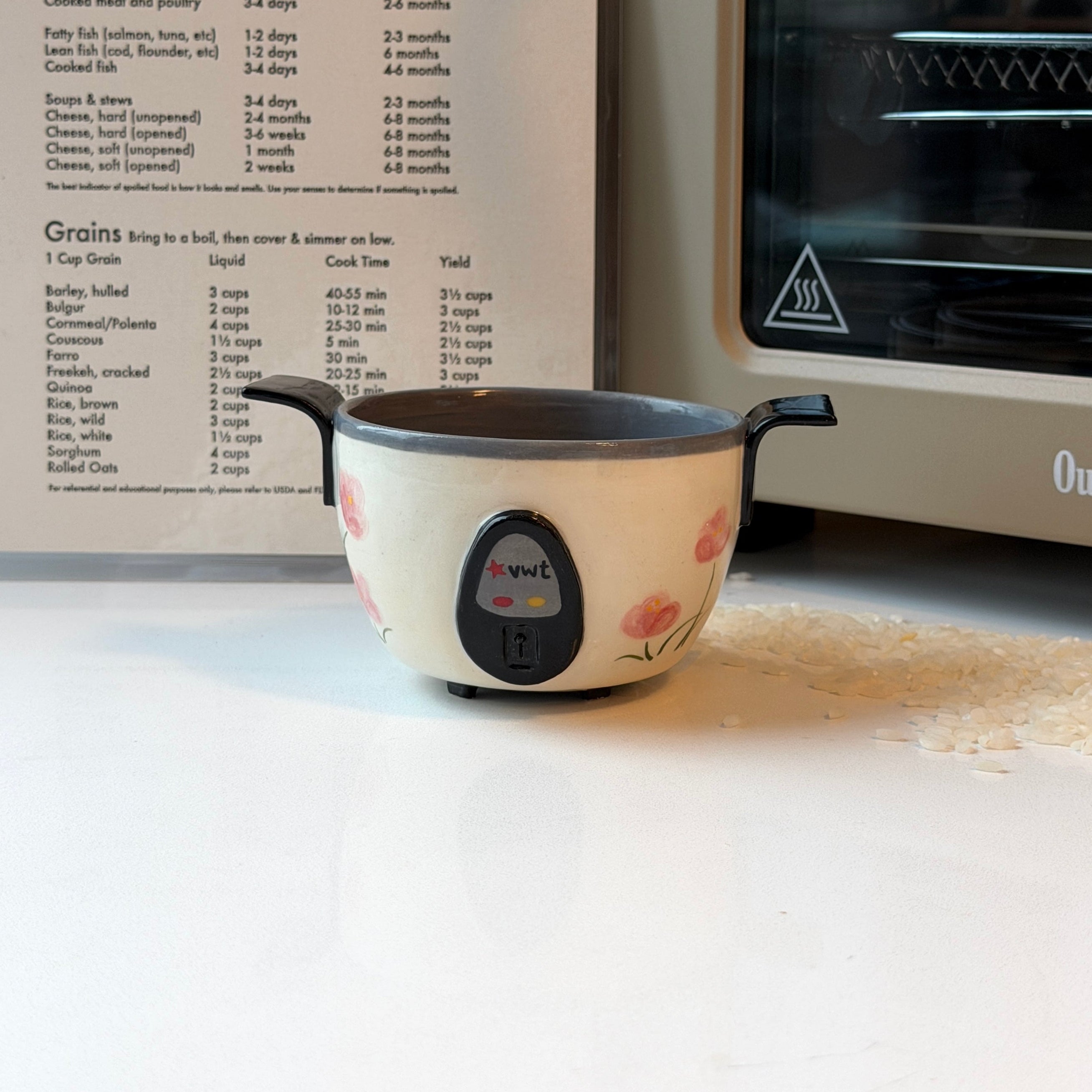 A small cream-colored ceramic bowl that looks like a rice cooker with pink floral patterns and black handles sits on a white countertop in a kitchen setting, with a recipe book and a toaster oven in the background.