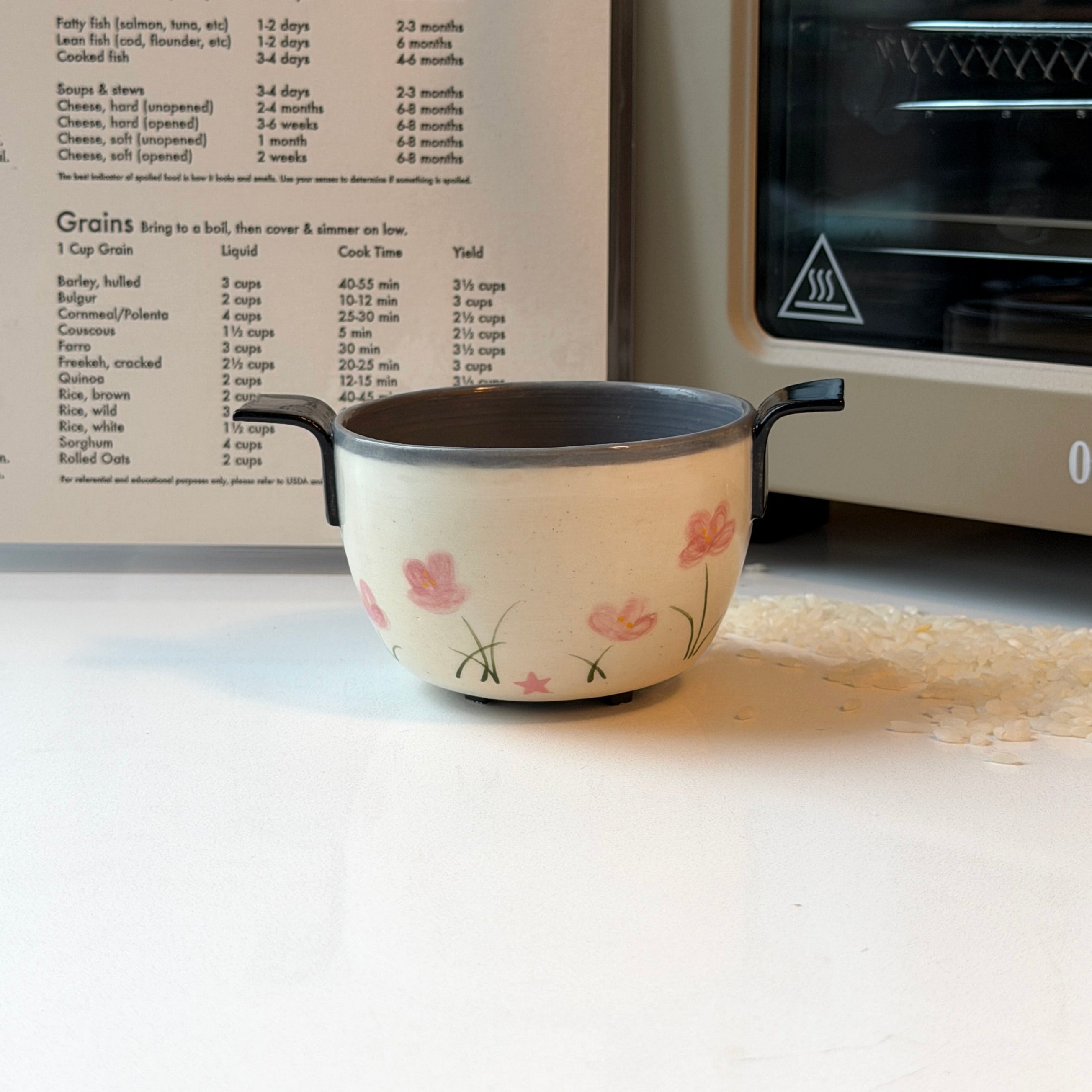 A small ceramic bowl that looks like a rice cooker with pink floral illustrations and black handles sits on a white countertop in a kitchen setting, with a recipe book and a microwave visible in the background.
