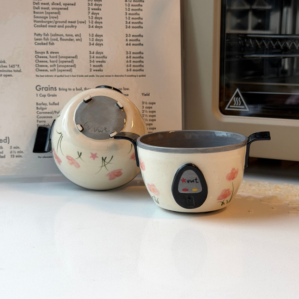 A cream-colored ceramic bowl that looks like a rice cooker with pink floral patterns and a black digital control display, shown next to a matching floral ceramic container on a white countertop, with a recipe book and a toaster oven in the background.