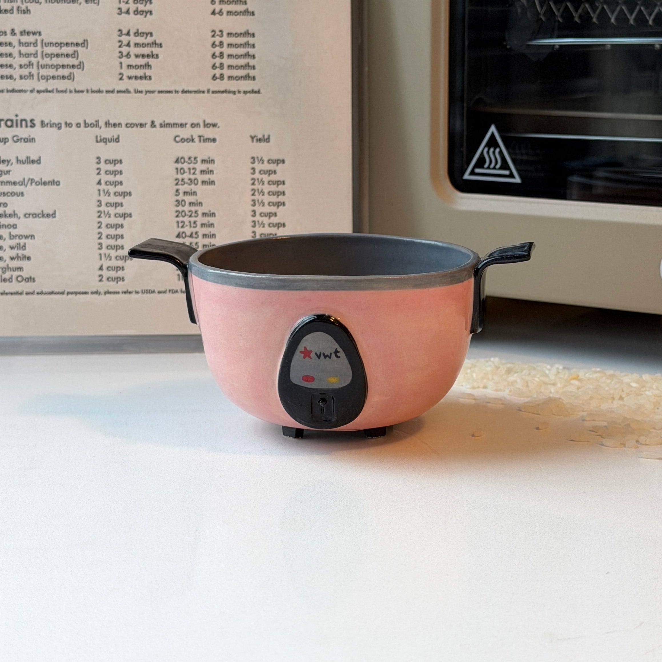 A small pink ceramic rice cooker with black handles sits on a white countertop in a kitchen setting, with a recipe book open behind it and a cream-colored oven to the right.