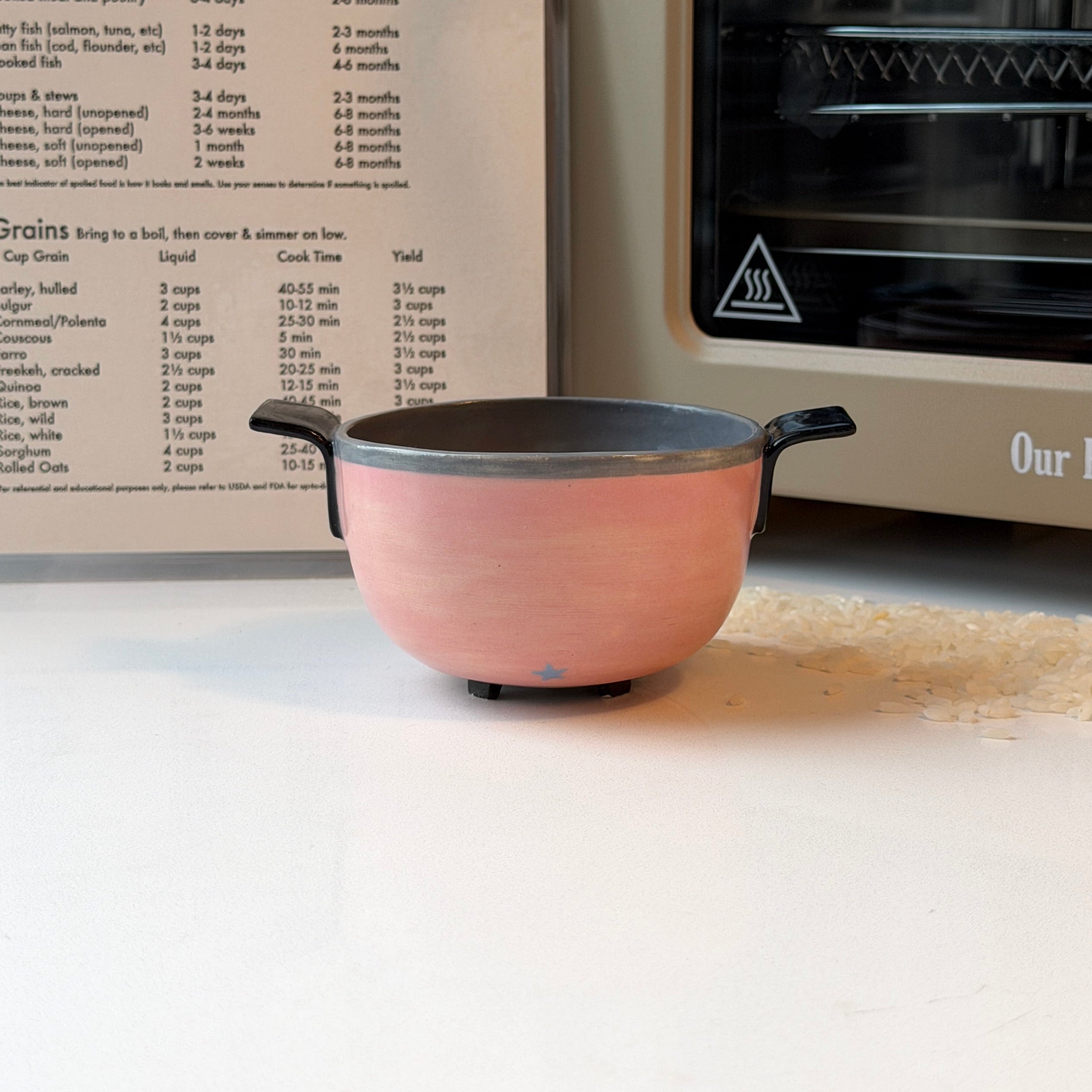A small blush pink ceramic rice cooker with black handles sits on a white surface in front of a cookbook and a small oven. The pot has a dark gray interior.