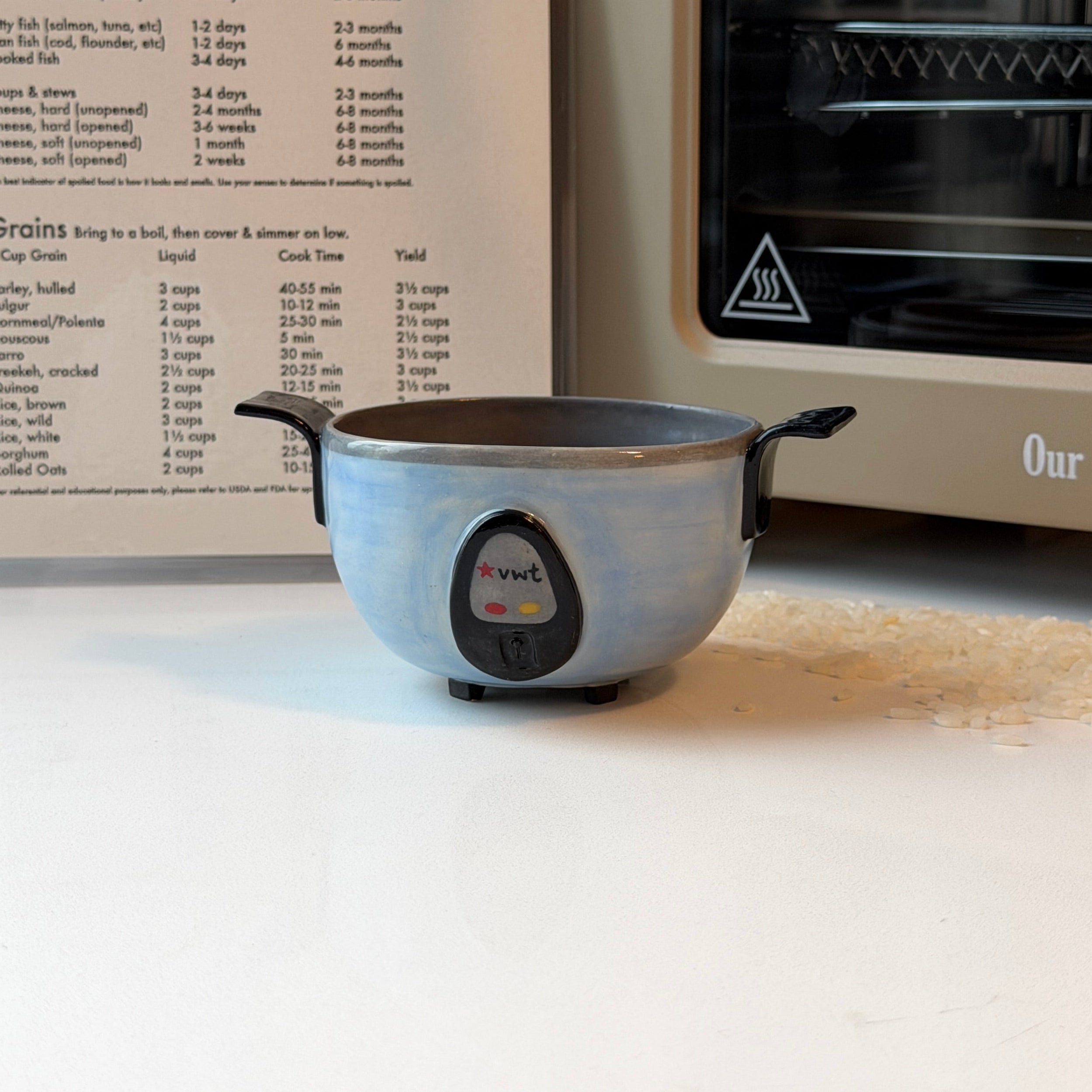 A small round ceramic rice cooker bowl with black handles sits on a white countertop in front of an open recipe book and a cream-colored toaster oven. 