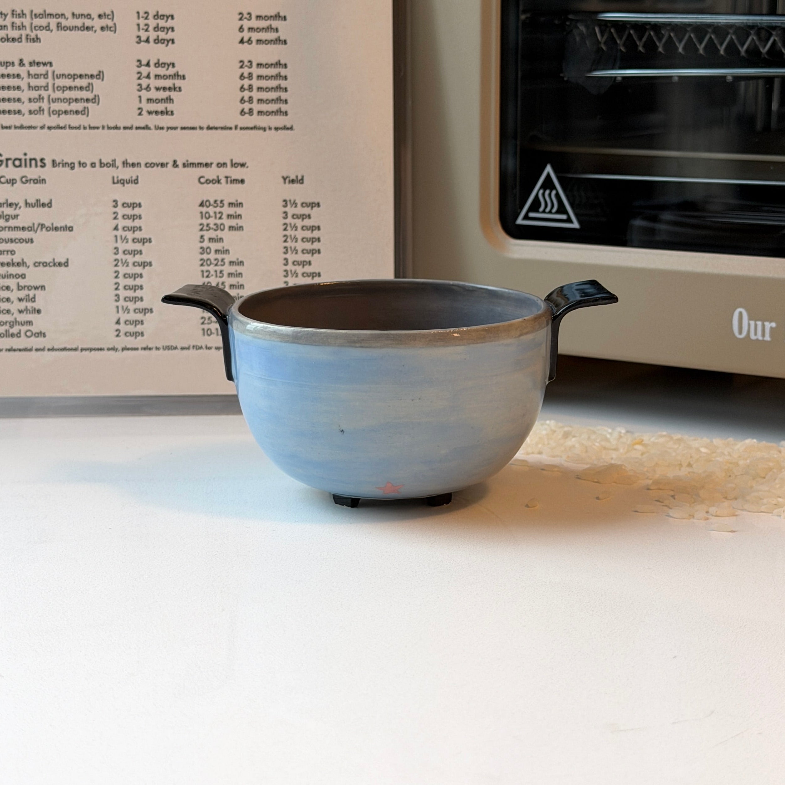 A small light blue ceramic bowl with black handles sits on a white surface in front of an open recipe book and a cream-colored oven. The bowl is the main product shown in a real kitchen-like setting.