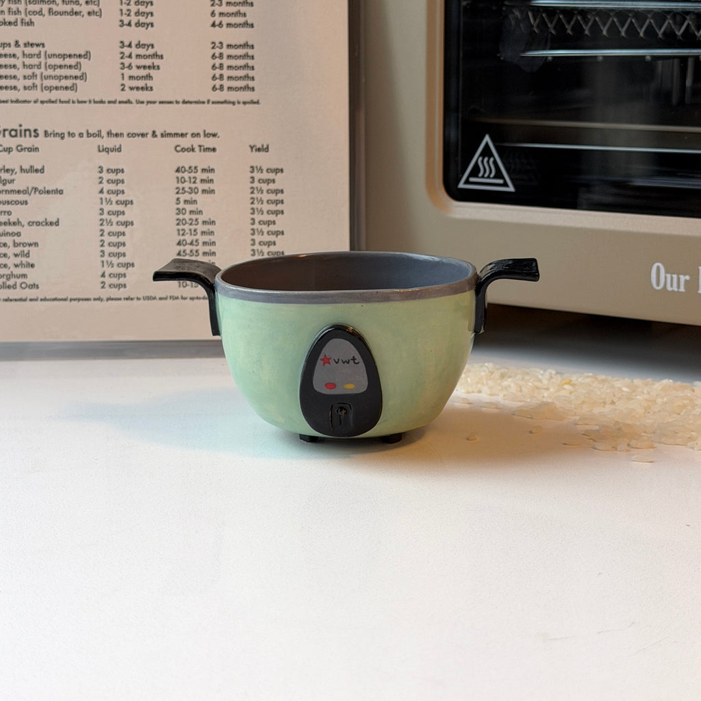 A small jade green ceramic bowl that looks like a rice cooker with black handles sits on a white countertop in a kitchen setting, with a recipe book and a cream-colored oven or toaster behind it.