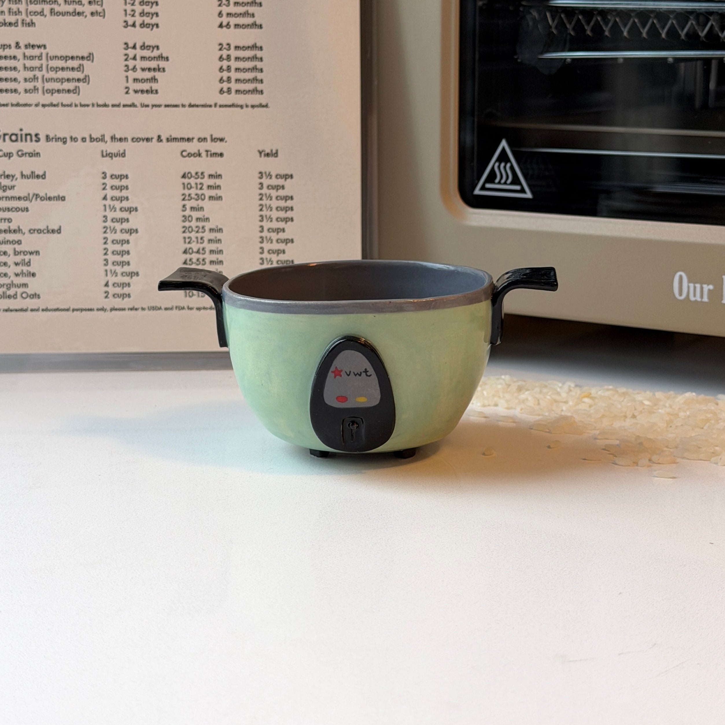 A small jade green ceramic bowl that looks like a rice cooker with black handles sits on a white countertop in a kitchen setting, with a recipe book and a cream-colored oven or toaster behind it.