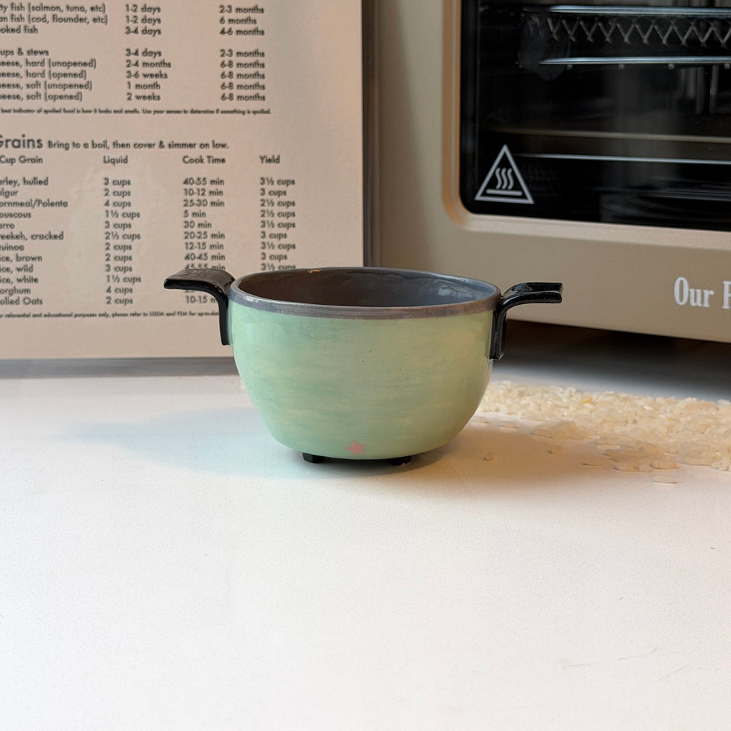A small handmade ceramic bowl that looks like a rice cooker with a light green glazed exterior and a dark gray interior sits on a white surface in front of an open recipe book and a beige oven.