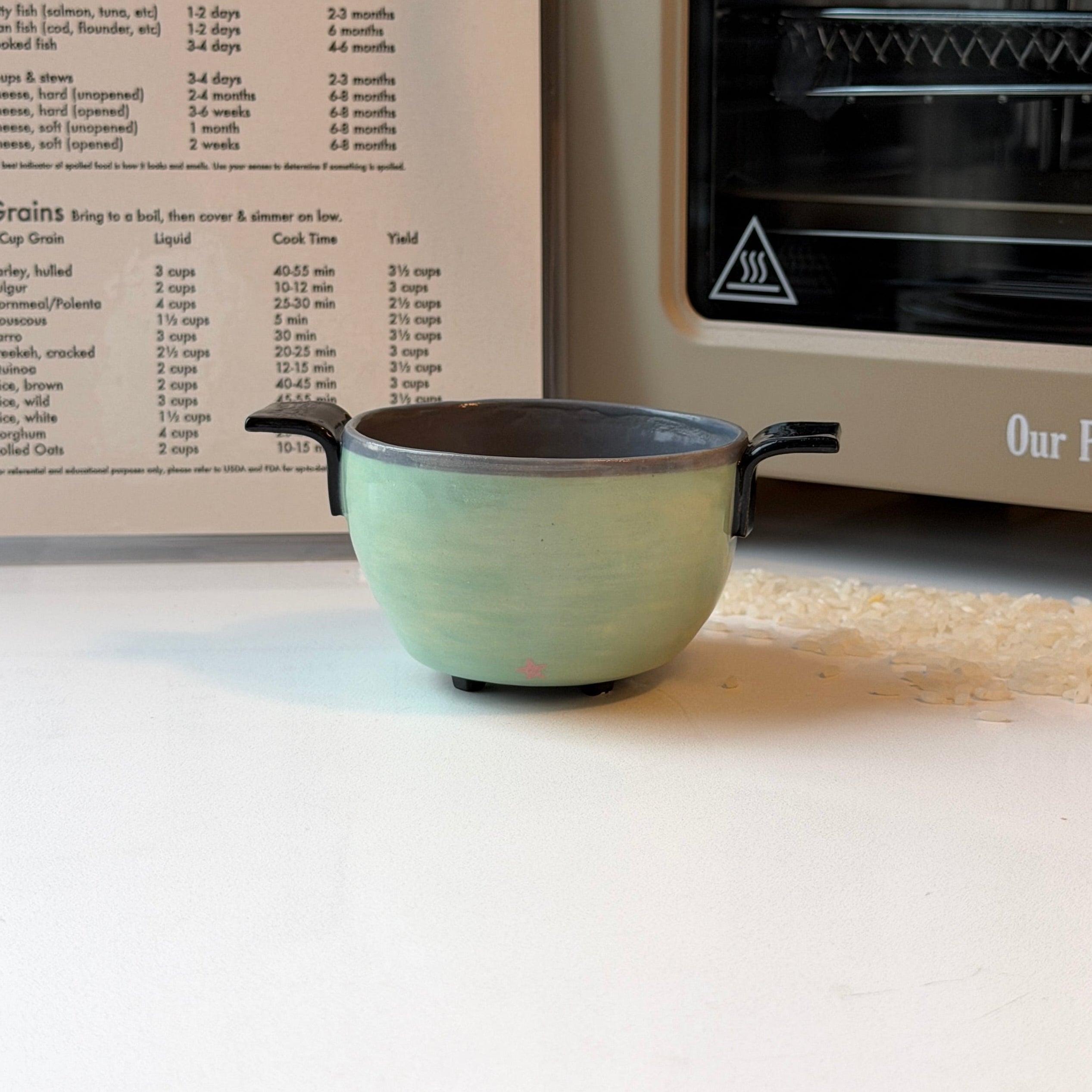 A small handmade ceramic bowl that looks like a rice cooker with a light green glazed exterior and a dark gray interior sits on a white surface in front of an open recipe book and a beige oven.