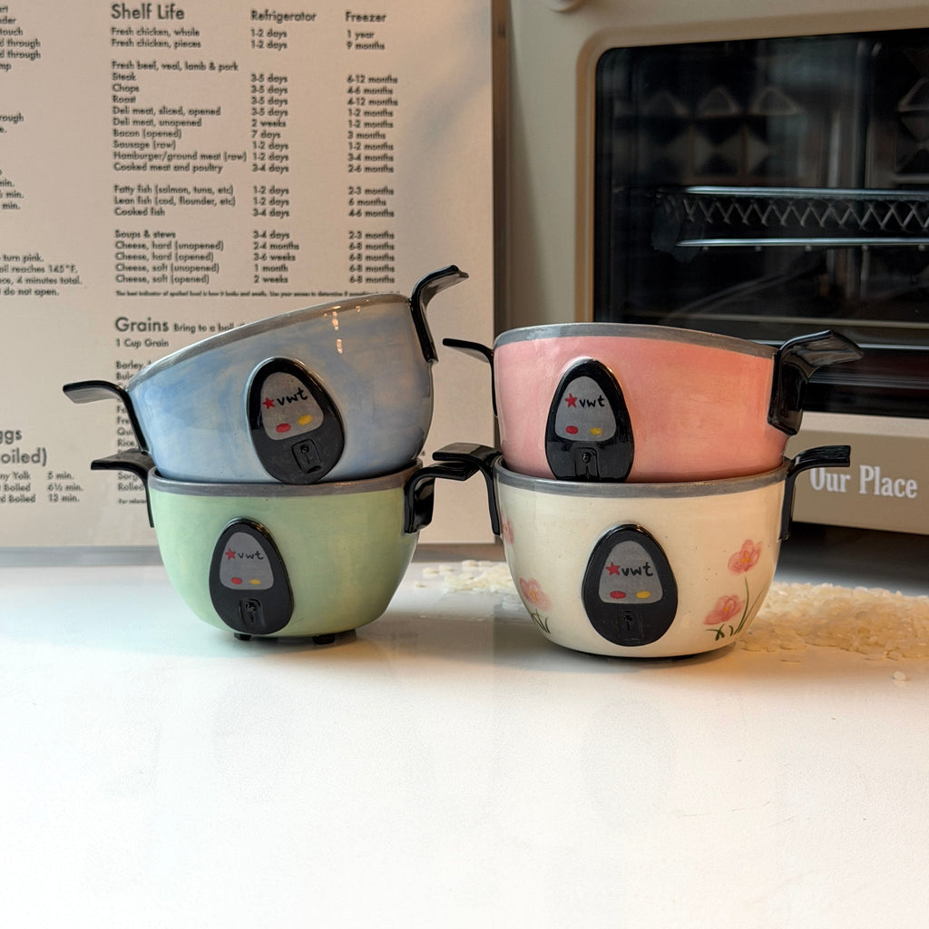 Four small ceramic bowls that look like rice cookers with black handles and a central logo, stacked in two pairs of two colors each: light blue, mint green, blush pink, and cream with pink floral details. They are arranged on a white countertop in front of a recipe book and a beige oven.
