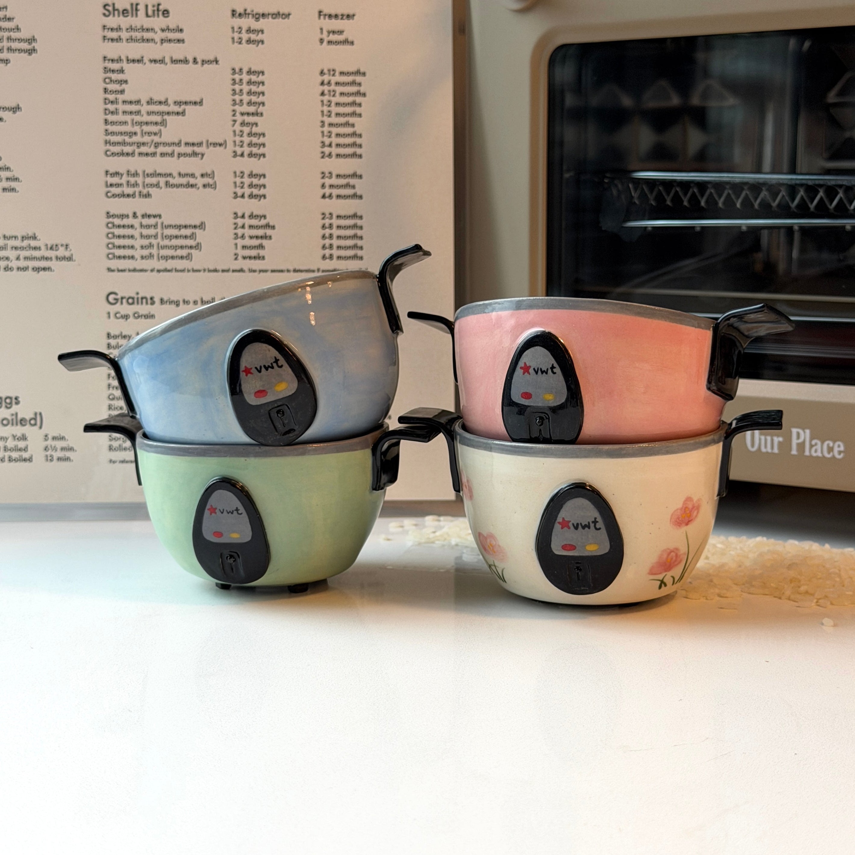 Four small ceramic bowls that look like rice cookers with black handles and a central logo, stacked in two pairs of two colors each: light blue, mint green, blush pink, and cream with pink floral details. They are arranged on a white countertop in front of a recipe book and a beige oven.